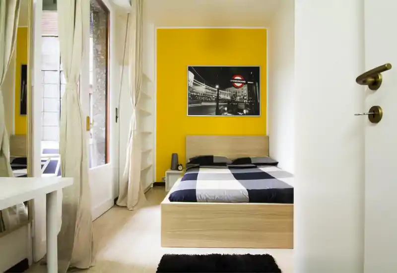 Bright, modern bedroom with a wooden double bed against a vivid yellow accent wall, large window with light curtains, and minimalist bedside table — clean, welcoming and well lit.