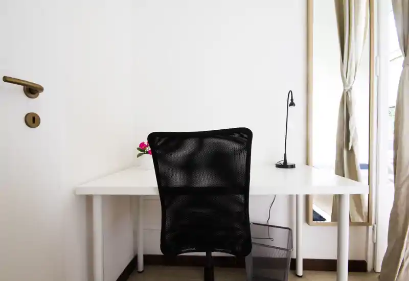 Bright minimalist workspace with a white desk, black mesh office chair, small desk lamp and a potted flower next to a sunlit window with light curtains.