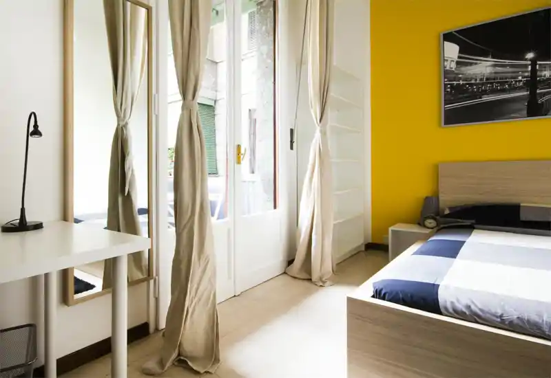 Bright, modern bedroom with a wooden bed frame, striped blue-gray bedding and a vivid yellow accent wall; large glass doors with neutral curtains provide abundant natural light.