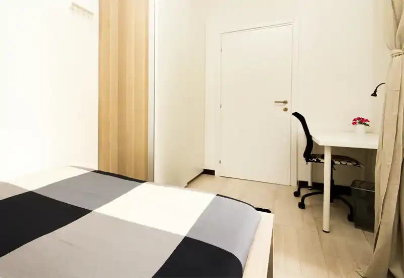 Bright, minimal bedroom with a large bed in the foreground, a wardrobe against the wall and a small desk with chair by the window — modern and tidy space ideal for a single occupant or short stays.