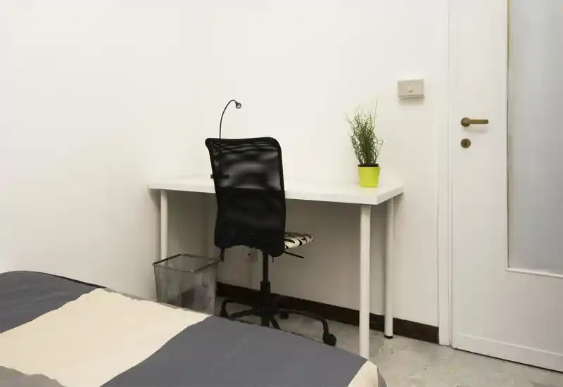 Rincón de dormitorio luminoso y minimalista con cama parcialmente visible y un escritorio blanco sencillo con silla ergonómica y pequeña planta — ideal como zona de estudio en una habitación compacta.