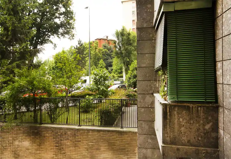 Vista de un área exterior/jardín junto a un edificio, con árboles, arbustos y un césped vallado visibles desde una perspectiva de planta baja/primer piso.