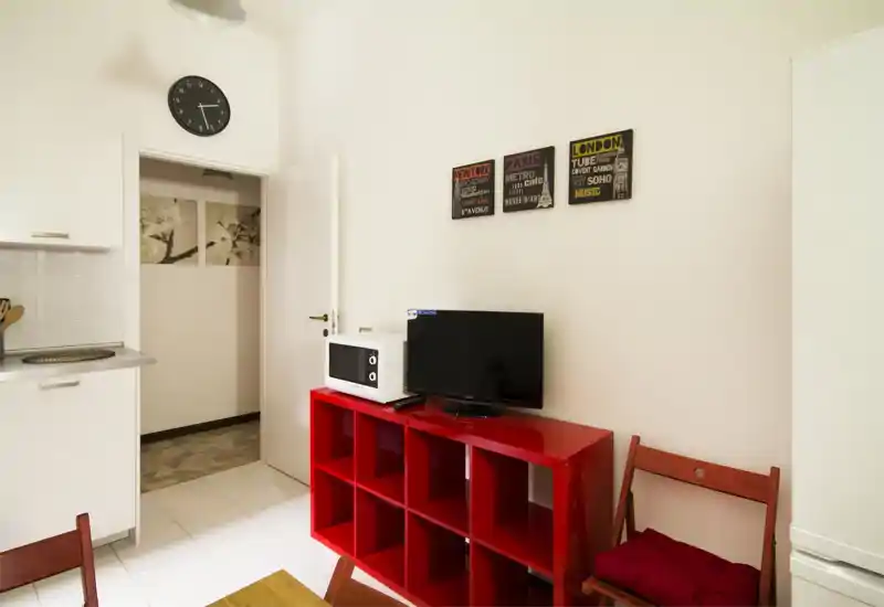 Zona de estar compacta y luminosa con unidad roja que sostiene la TV y el microondas, sillas de madera simples y paredes claras para un estilo moderno y limpio.