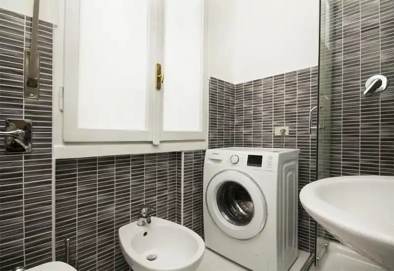 Baño compacto con azulejos mosaico grises, bidé, lavabo, mampara de ducha y lavadora de carga frontal junto al lavabo.