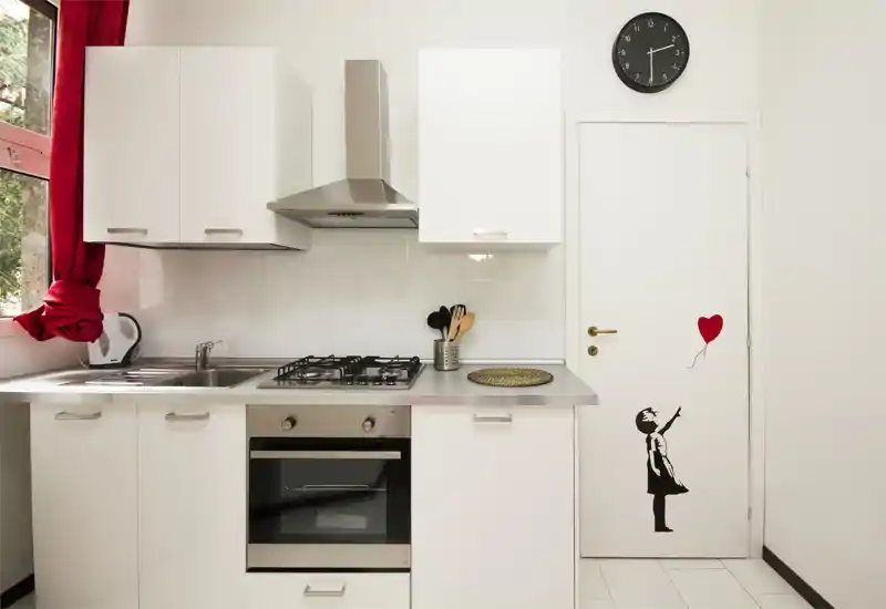 Cocina moderna y compacta con armarios blancos, encimera de acero inoxidable, placa de gas y horno; luz natural y una cortina roja que añade un toque animado.