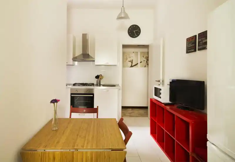 Cocina de concepto abierto, luminosa y ordenada, con horno empotrado, placa de gas y muebles blancos, combinada con una mesa de madera y estantería roja.