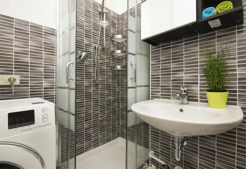 Baño moderno y compacto con ducha de esquina de vidrio, lavabo de pared y lavadora; azulejos mosaico grises horizontales y estanterías abiertas para artículos de aseo.