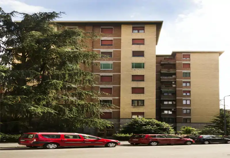 Vista exterior de un edificio residencial de altura media con coches aparcados y árboles frente a la calle; adecuada para mostrar la fachada y el entorno.