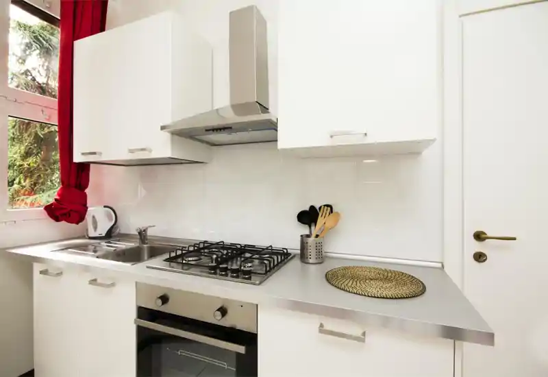 Cocina moderna y compacta con encimera de acero inoxidable, placa de gas y horno empotrado; mucha luz natural desde la ventana y detalles cálidos como la cortina roja y el recipiente de utensilios.
