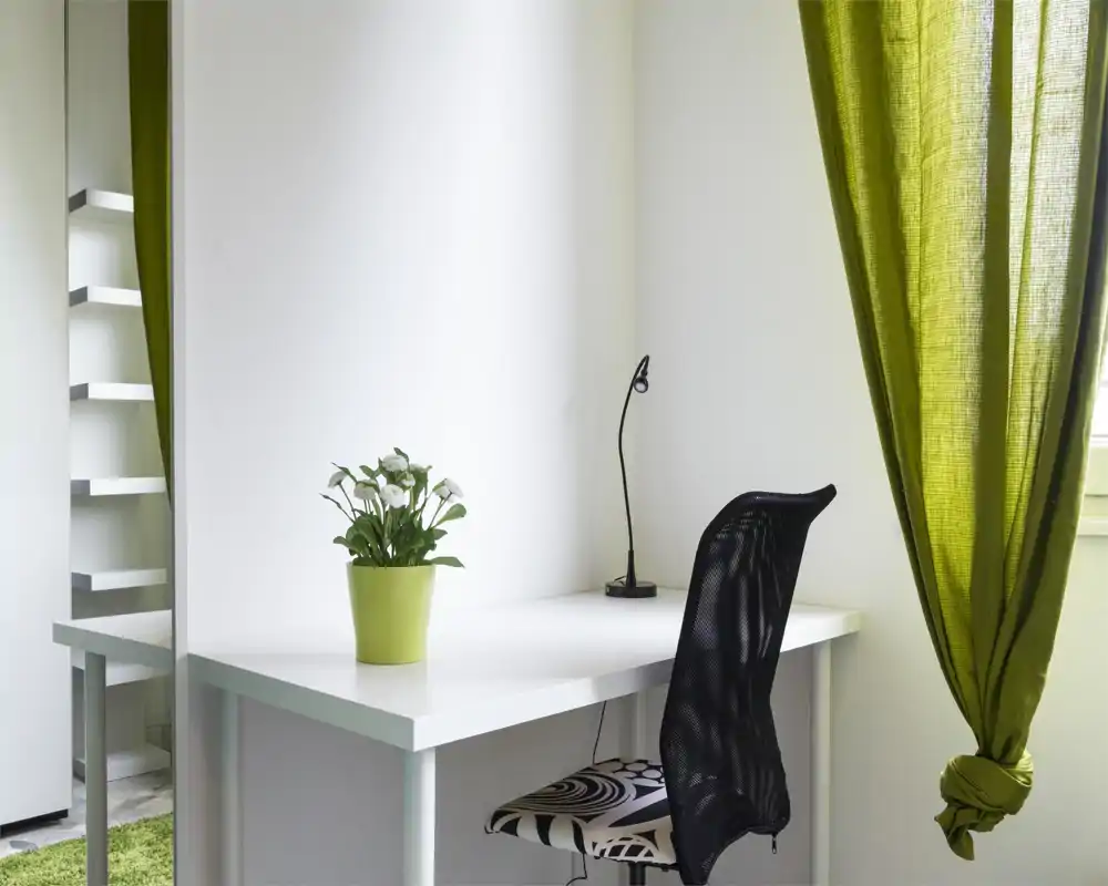 Rincón luminoso y minimalista del salón con escritorio blanco, silla moderna de malla y detalles verdes como una planta en maceta y cortina a juego.
