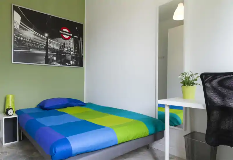 Dormitorio moderno y luminoso con cama doble con ropa de cama azul y verde, mesita con reloj y lámpara, cuadro en la pared y un escritorio compacto con silla — ideal para estudiante o persona sola.