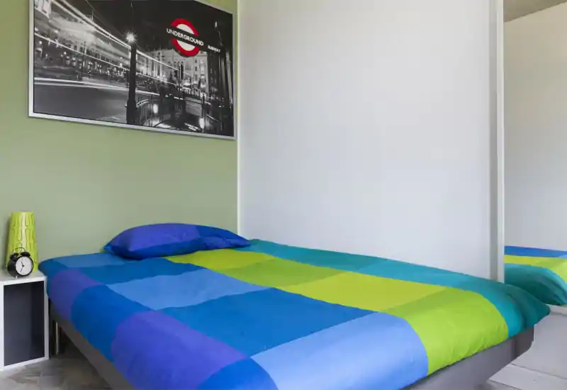 Dormitorio luminoso y ordenado con una cama doble de colores, mesita con lámpara y un cuadro en la pared. Estilo moderno y buena luz natural que lo hace acogedor.