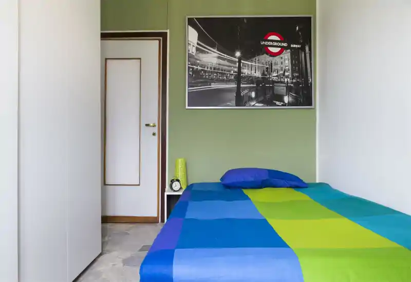 Dormitorio individual luminoso y ordenado con edredón azul y verde, almohada y mesita con despertador; un cuadro en blanco y negro sobre pared verde claro añade personalidad.