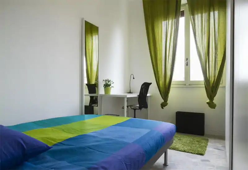 Dormitorio luminoso y ordenado con una cama doble colorida, cortinas verdes y una pequeña zona de trabajo junto a la ventana — ideal para una estancia cómoda y funcional.