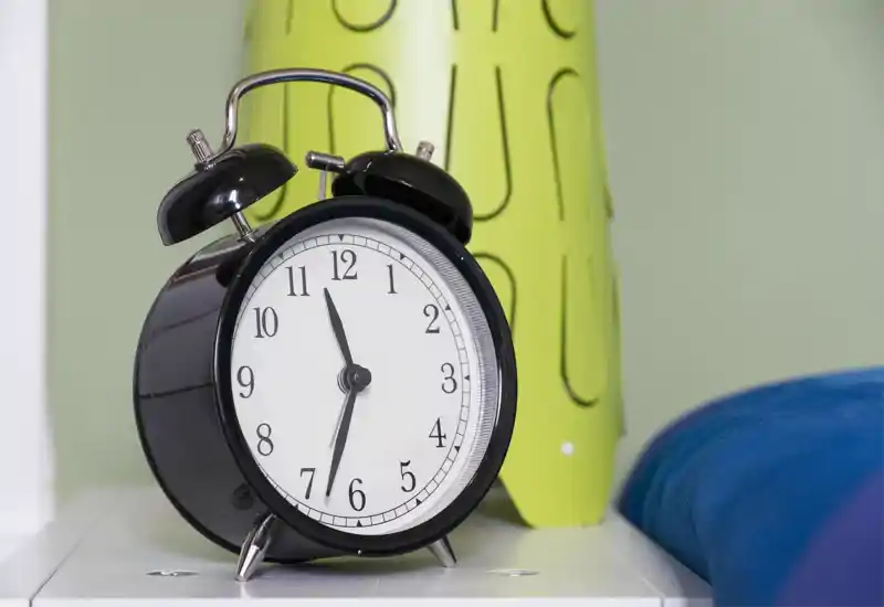 Primer plano de un despertador sobre la mesita de noche con un atisbo de la ropa de cama en el borde; imagen nítida y luminosa, ideal para mostrar detalles del mesita.