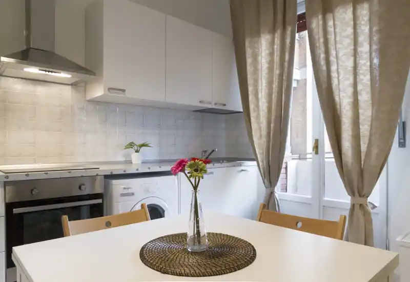 Cocina compacta y luminosa con muebles blancos, horno integrado y lavadora, mesa pequeña con flores y cortinas hasta el suelo que dan a un balcón/puerta.