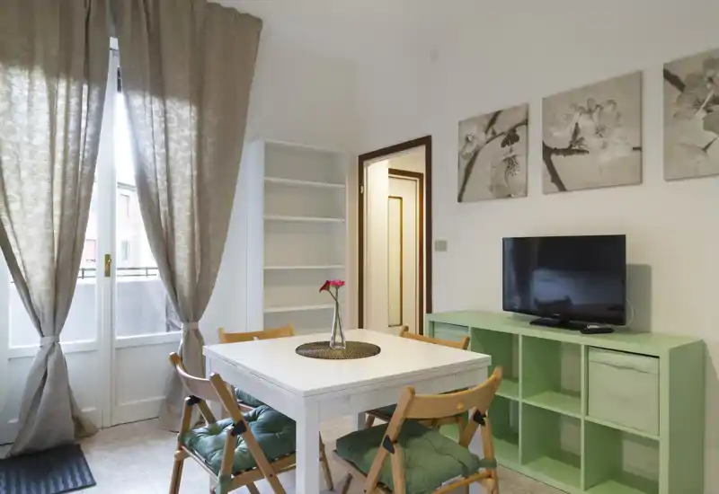 Zona de estar/comedor luminosa y bien decorada con mesa blanca central, sillas plegables acolchadas, cuadros decorativos y televisión sobre un mueble verde menta. Amplias ventanas con cortinas proporcionan luz natural y ambiente acogedor.