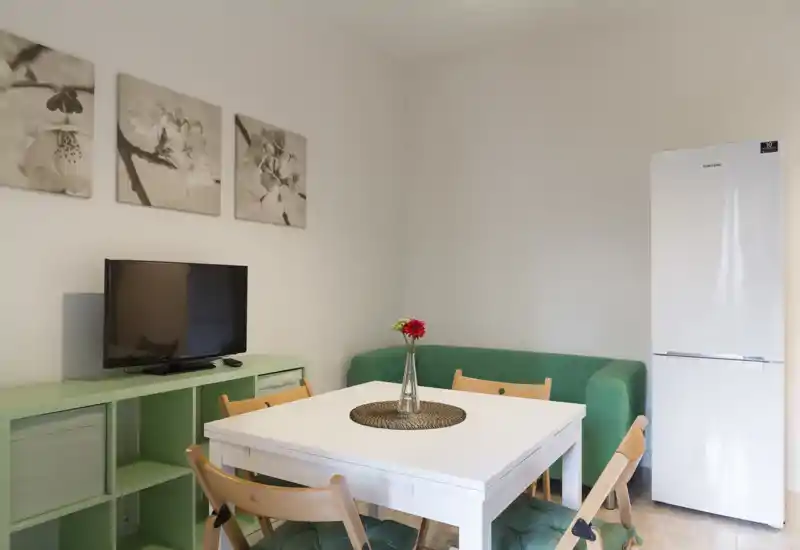 Zona de estar compacta y luminosa con un sofá verde pequeño, mesa blanca con sillas de madera y televisor sobre un mueble verde menta — perfecta para un estudio acogedor.