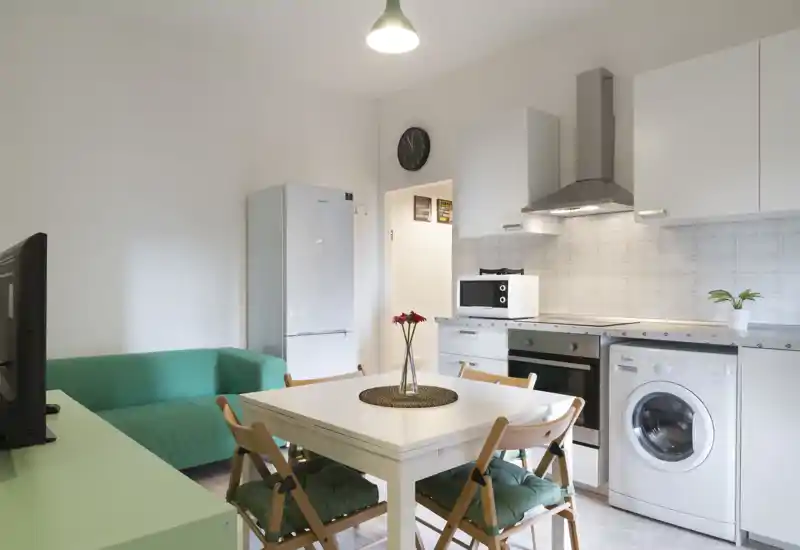 Cocina abierta moderna y luminosa con mobiliario blanco, horno, lavadora y mesa de comedor para cuatro. Espacio limpio y sencillo con un pequeño sofá y flores frescas.