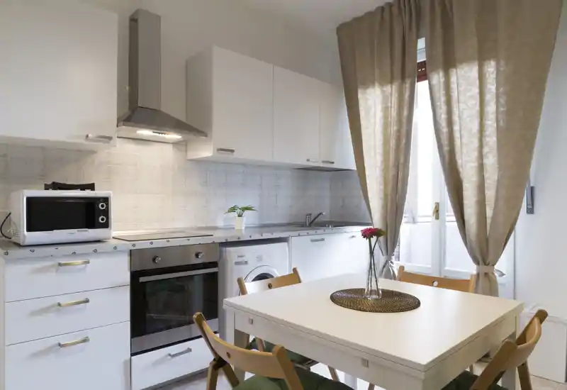 Cocina pequeña y luminosa con muebles blancos, horno integrado y lavadora, y una mesa de comedor para cuatro junto a la ventana.