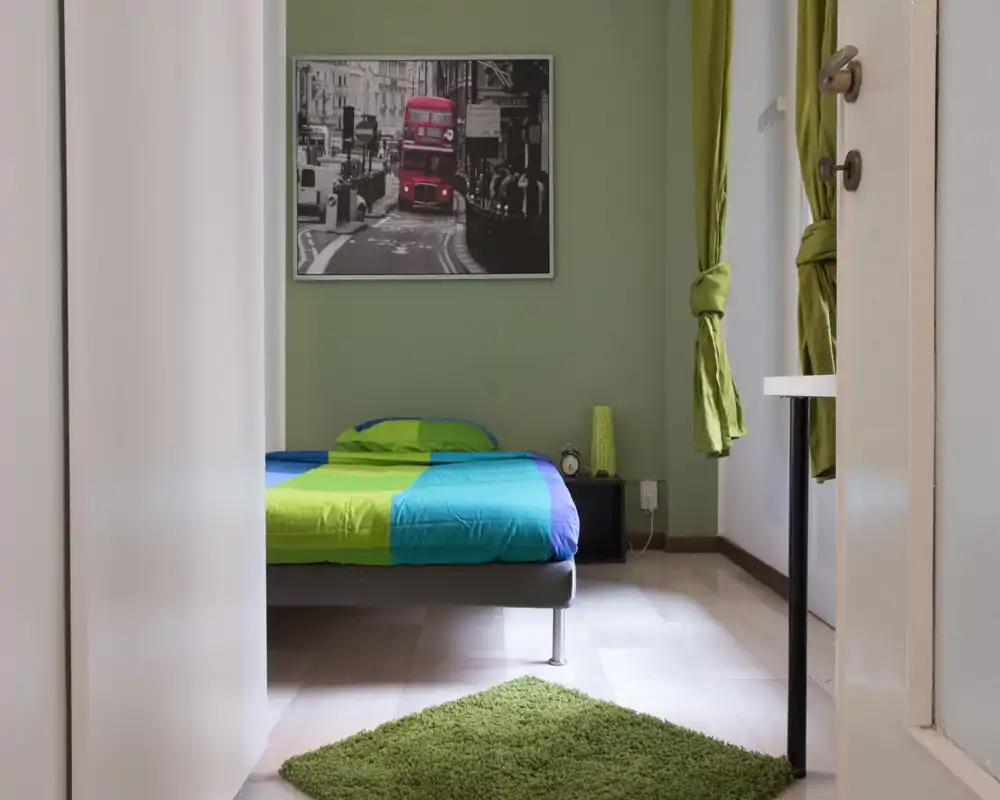Dormitorio luminoso y sencillo con cama doble vestida con ropa de cama verde y azul, mesita de noche y una foto enmarcada en la pared. La luz natural, las cortinas verdes a juego y una alfombra pequeña crean un ambiente fresco y acogedor.