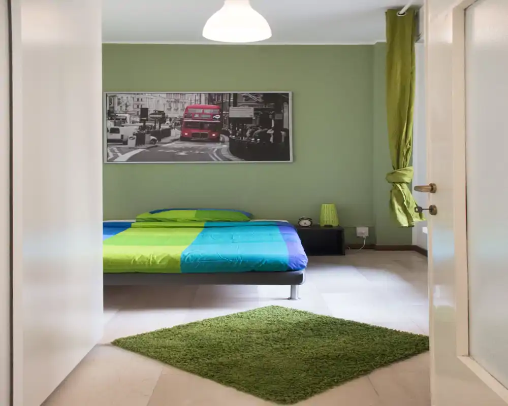 Dormitorio moderno y luminoso con cama baja de ropa de cama verde y azul, alfombra verde y cuadro a juego; líneas limpias y buena luz natural.