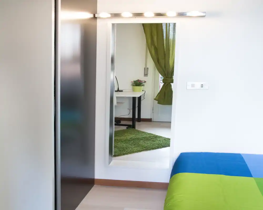 Rincón de dormitorio moderno y luminoso con ropa de cama colorida y un gran espejo que muestra un espacio de trabajo contiguo con alfombra verde.