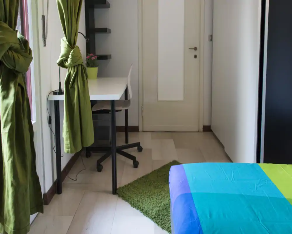 Dormitorio individual luminoso y ordenado con colcha colorida, alfombra verde y un pequeño escritorio junto a la ventana — ideal para estudiante o persona sola.