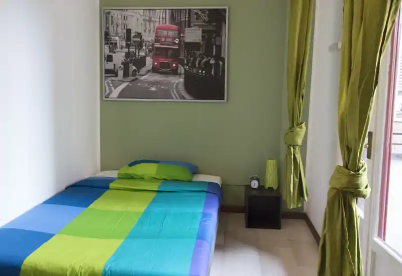Dormitorio pequeño y luminoso con una cama colorida, mesita cúbica y luz natural procedente de una ventana/puerta. Decoración sencilla y moderna con una foto de la ciudad encima de la cama.
