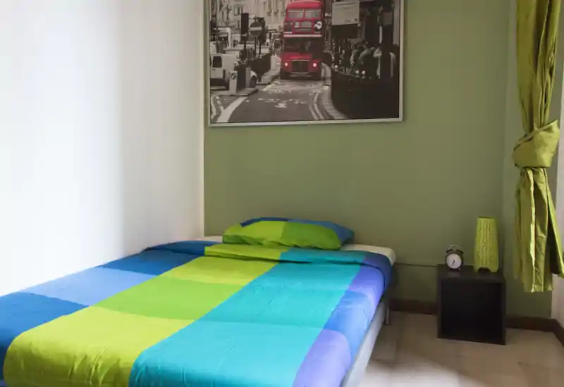 Dormitorio luminoso y bien presentado con una cama colorida, mesita con despertador y una imagen de la ciudad sobre la cabecera.