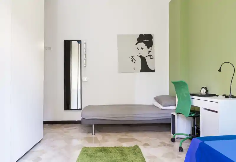 Camera da letto semplice e moderna con letto singolo basso, scrivania e parete verde. Spazio luminoso e ordinato ideale per uno studente o una singola persona.