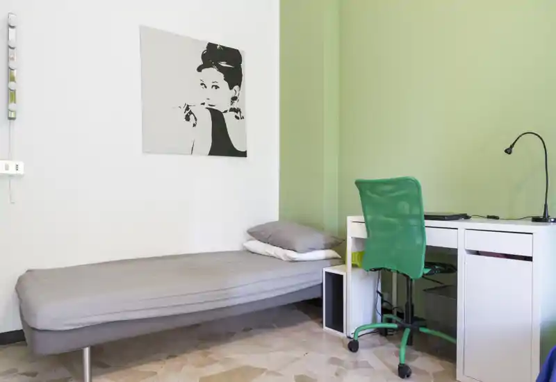 Camera singola semplice e luminosa con letto ben sistemato, una piccola mensola e una scrivania bianca con sedia verde — ideale per uno studente o un occupante singolo.