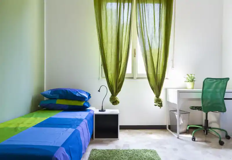 Camera da letto singola luminosa e ordinata con letto rifatto, piccolo comodino e una scrivania vicino alla finestra. Accenti verdi (tende, tappeto, sedia) creano un aspetto fresco e coordinato, ideale per studenti o affitti brevi.