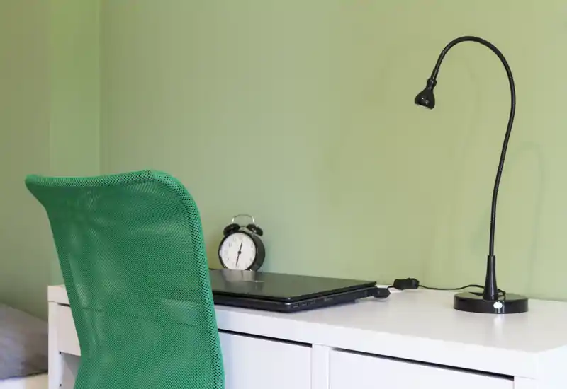Compact bedroom workspace with a white desk, green mesh chair, bedside corner of the bed visible, a closed laptop, alarm clock and an adjustable desk lamp — bright and tidy.