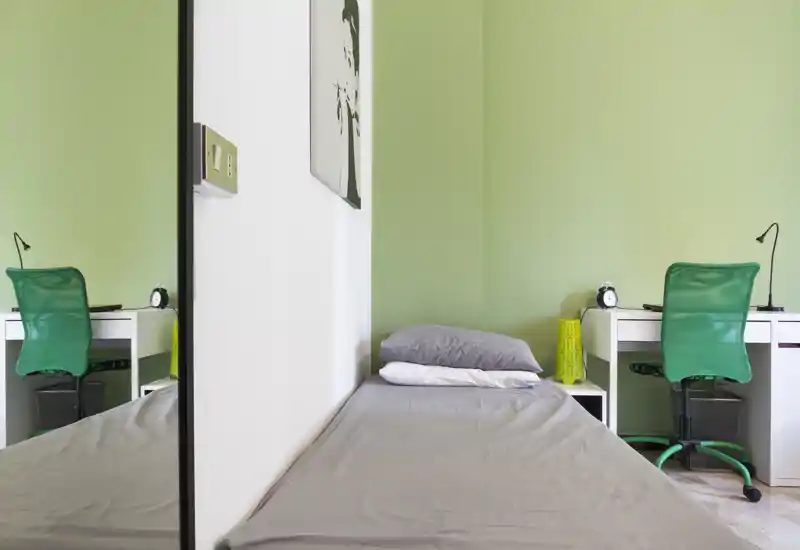 Camera da letto compatta e luminosa con un singolo letto contro una parete verde chiaro, comodino e scrivania con sedia verde: arredo moderno, pulito e funzionale per uno studente o un ospite singolo.