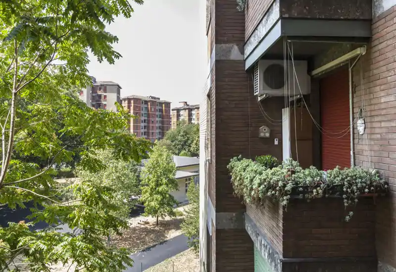 Small urban balcony with potted succulents, a closed red shutter and an external air-conditioning unit; leafy trees and apartment blocks are visible beyond, offering a pleasant city-facing outdoor spot.