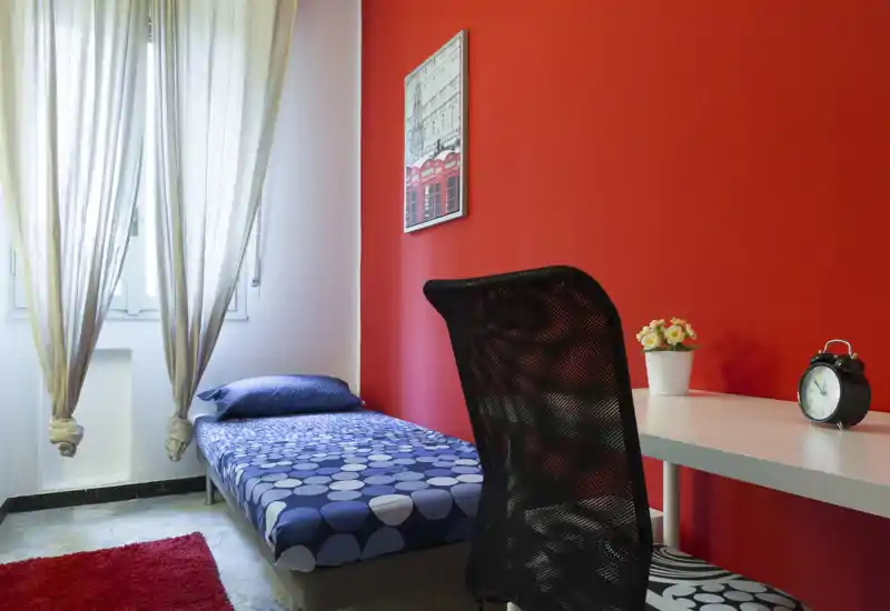 Dormitorio compacto con cama individual junto a una ventana luminosa, ropa de cama azul estampada, escritorio blanco con despertador y una silla negra de malla frente a una pared roja.