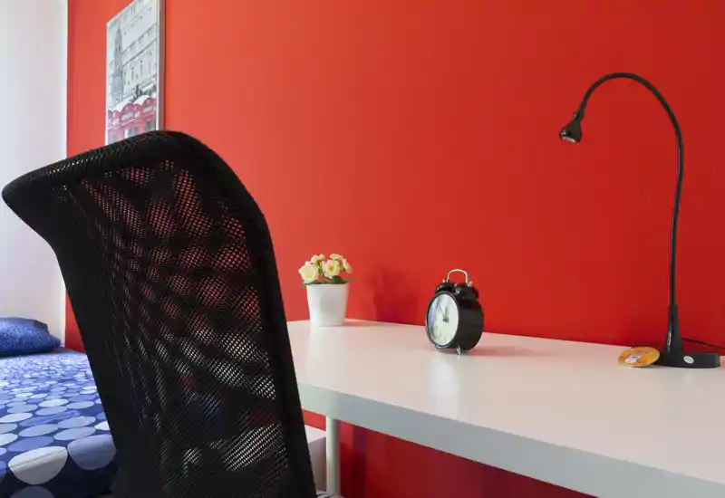Rincón de dormitorio luminoso y moderno con escritorio blanco frente a una pared roja intensa, silla negra de malla, despertador y un vistazo a la cama azul con estampado.