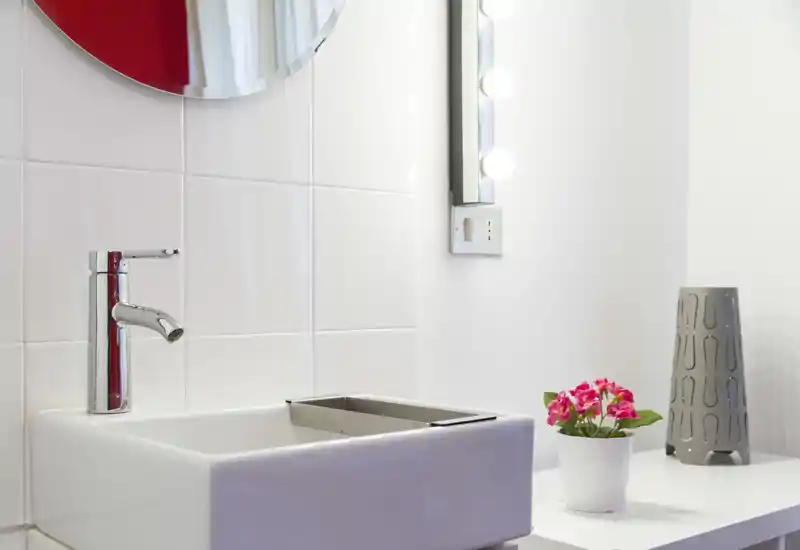 Modern bathroom vanity with a white rectangular sink, chrome faucet, round mirror and illuminated vanity lights; small potted flowers add a decorative touch.