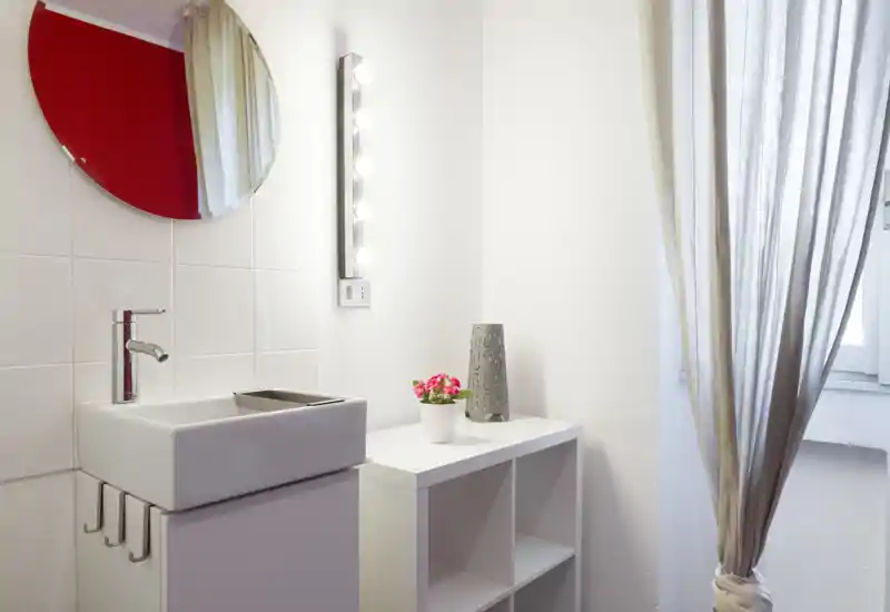 Small, bright bathroom corner with a compact sink, round mirror and shelving unit next to a curtained window. Simple, modern styling with clean tiles and decorative plant.