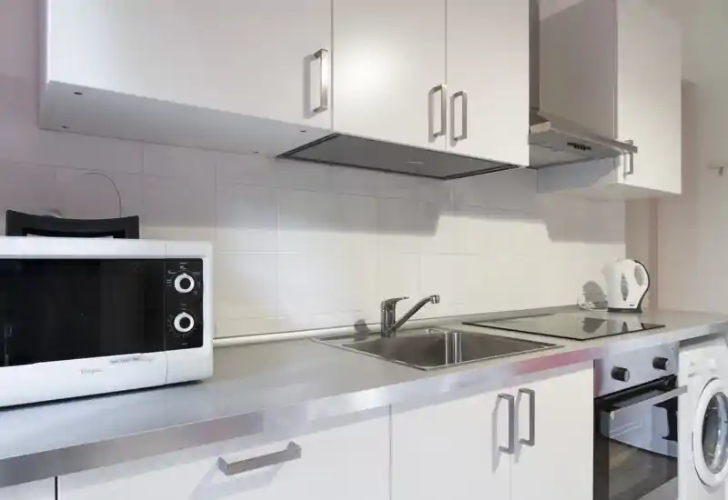 Modern compact kitchen with stainless countertop, built-in sink, electric hob, oven and microwave. Bright white cabinets and tidy layout ideal for short-term rentals.