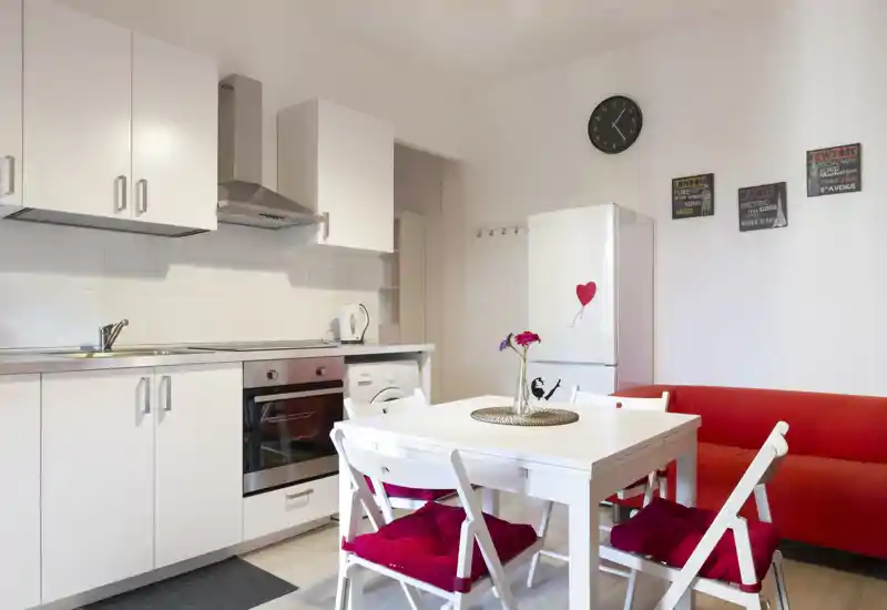 Bright modern kitchen with white cabinetry, integrated oven and a dining table with red-cushioned chairs; a small red sofa adds cozy seating and decorative touches.