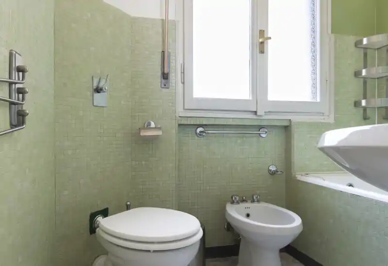 Baño compacto con azulejos mosaico verdes, inodoro, bidé y bañera parcialmente visible junto a un lavabo blanco; luz natural entra por una ventana esmerilada.