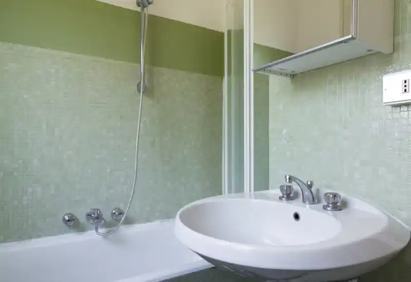 Clean bathroom view with a white sink and bathtub, green mosaic tiled walls and a handheld showerhead — bright and functional fixtures.