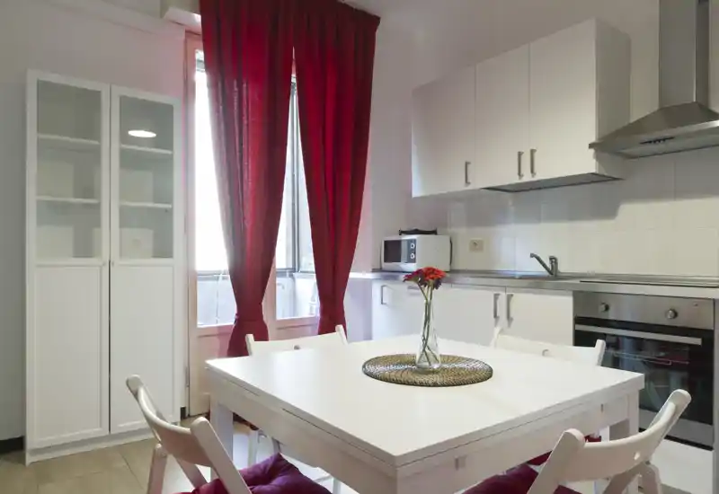 Modern fitted kitchen with a white dining table in the foreground, red curtains and a vase of flowers creating a warm, inviting atmosphere. Stainless steel oven and hood, microwave and wall cabinets provide practical storage and appliances.