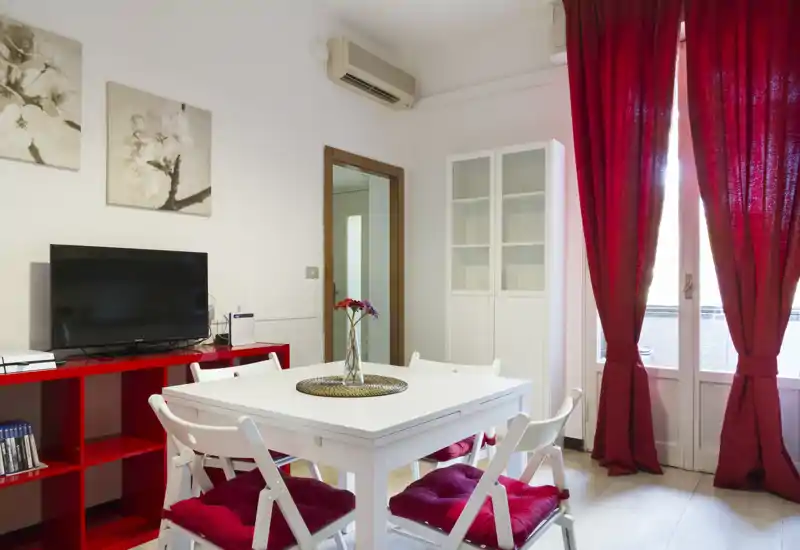 Bright, modern dining area with a white table, red-cushioned chairs and a glass-door balcony; tasteful red accents and a TV unit create a cozy, functional space.