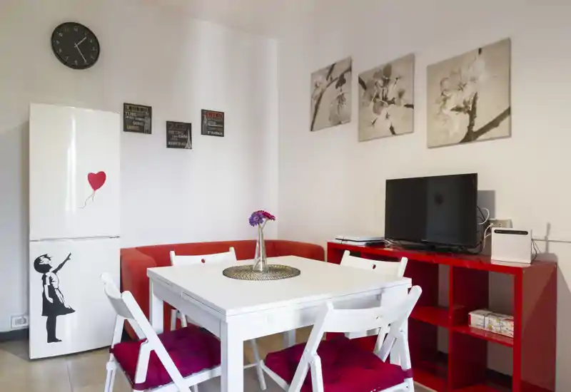 Bright, modern dining corner with a white table, four chairs with red cushions, a red side unit holding a TV and decorative wall art — comfortable and well styled for meals.