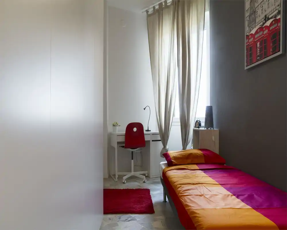 Bright, tidy single bedroom with a colorful orange-and-magenta bedspread, a bedside shelf and a small desk by the window offering natural light.