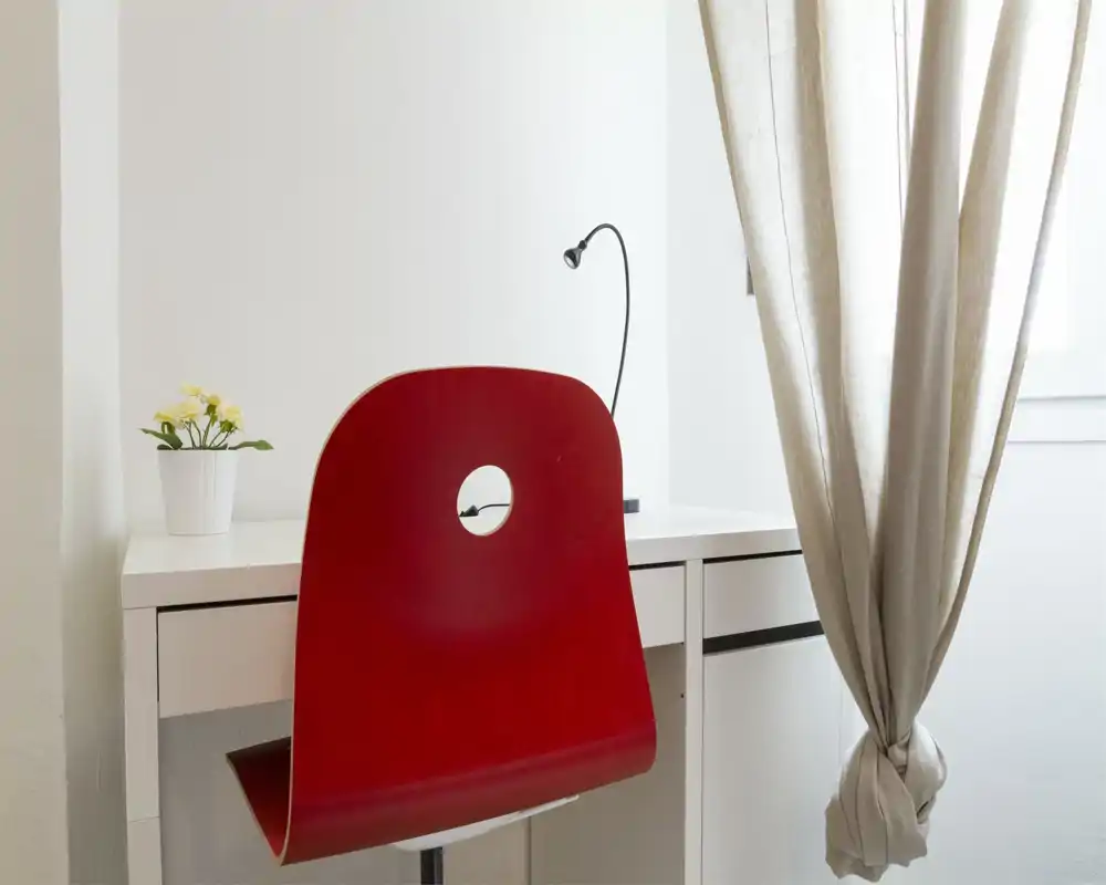 Bright, minimal living/work corner with a red chair, white desk, task lamp and a small potted plant by a curtained window.