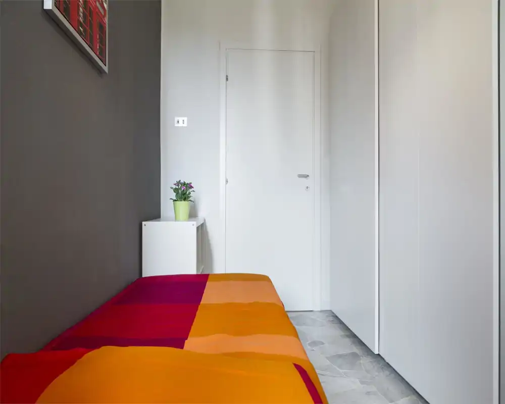 Compact, tidy bedroom with a colorful orange and red bedspread, a white nightstand and a potted plant; neutral walls and built-in wardrobe create a bright, modern feel ideal for short-term rentals.